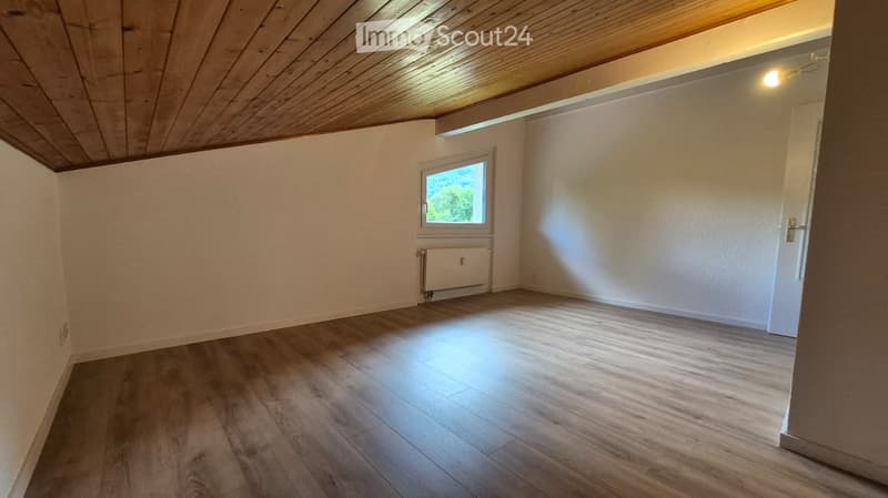 Empty room with wooden flooring, white walls, and a single window letting in natural light. The room features a radiator below the window and a wooden ceiling with recessed lighting.
