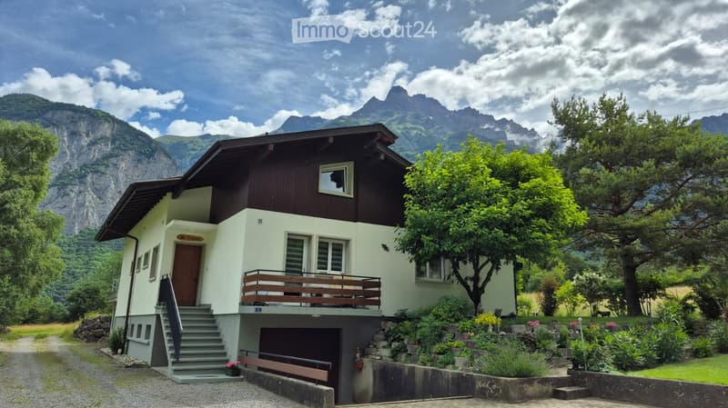 Two-story house with a balcony, garage, and surrounded by greenery and mountains.