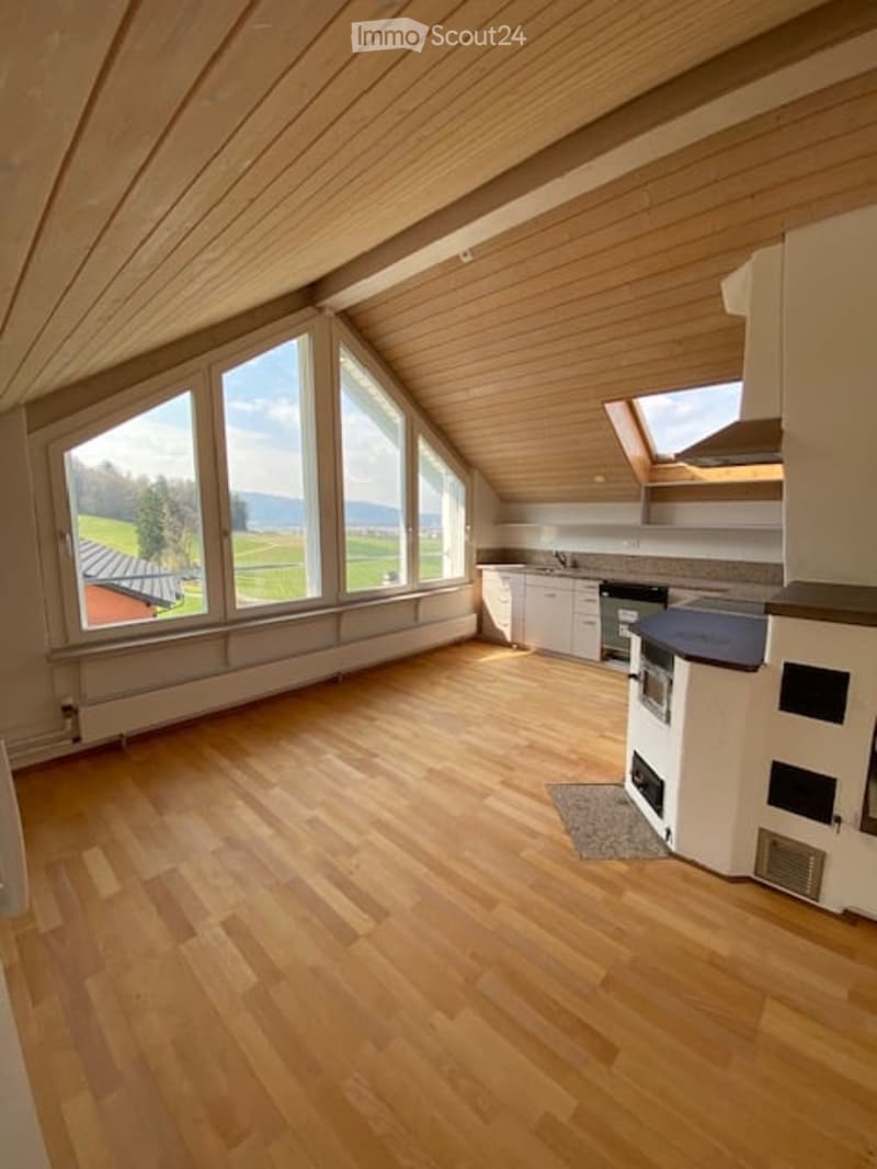 Bright, open-plan attic space with wooden floors, large windows offering scenic views, and a modern kitchen area with white cabinets, stainless steel appliances, and a black countertop.