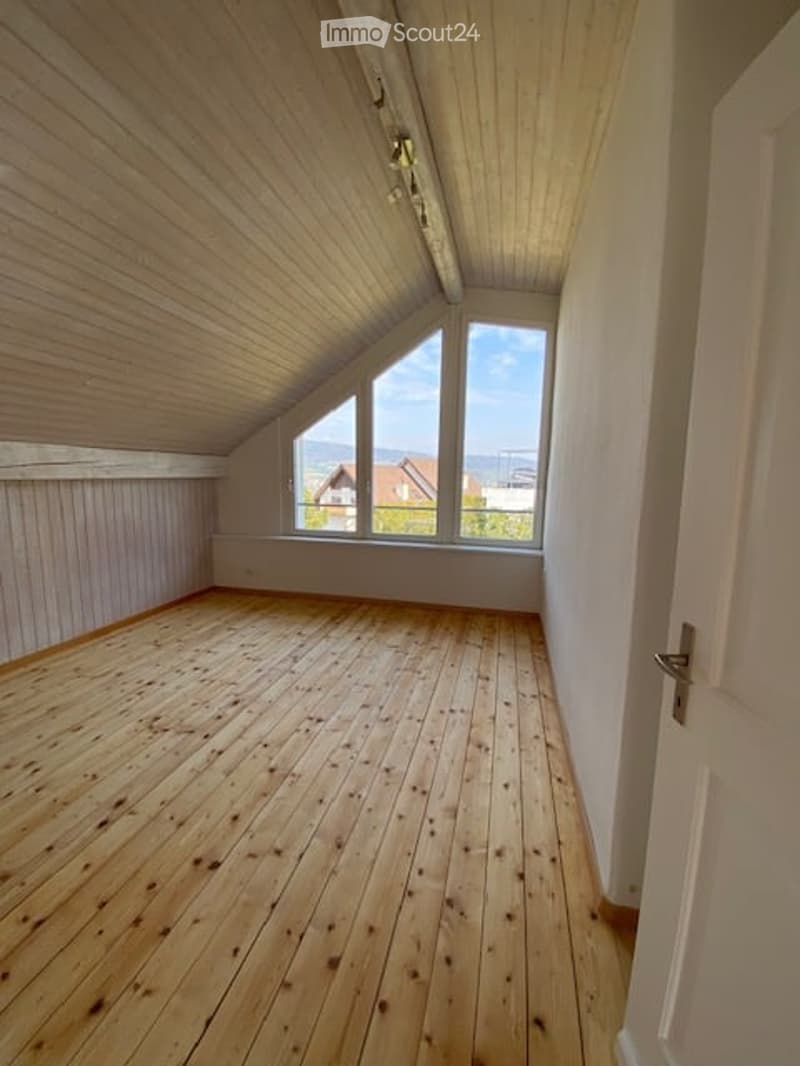 Empty attic room with wooden floors and slanted wooden ceiling, large windows offering a view of the landscape.