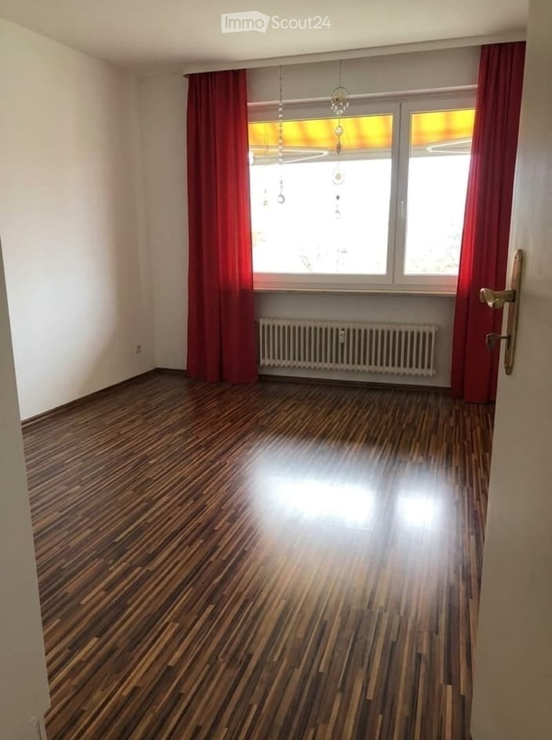 Empty room with wooden flooring, large windows with red curtains, radiator, and a door with a golden handle.