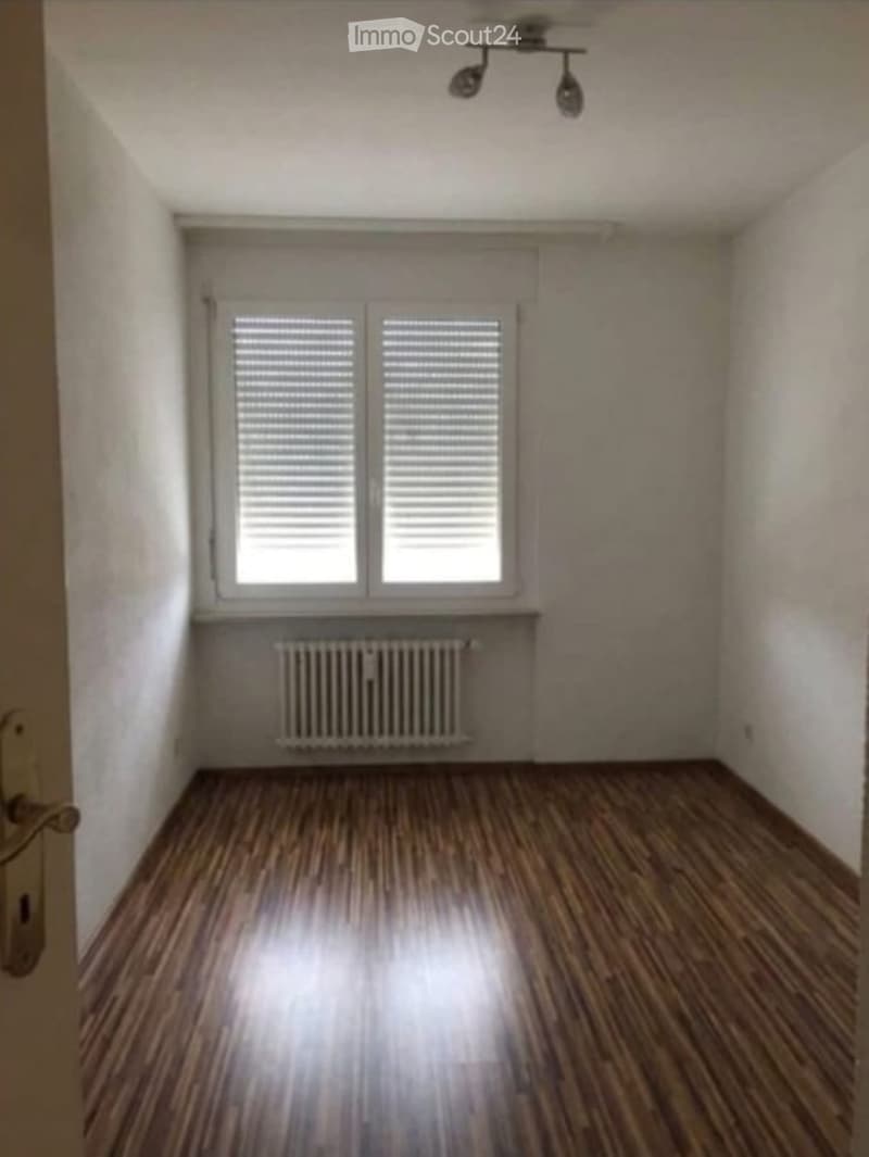 Empty room with wooden flooring, white walls, a window with blinds, a radiator, and a ceiling light fixture.