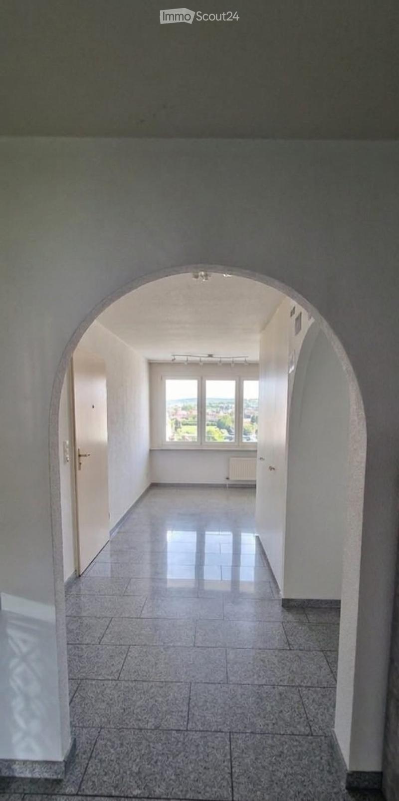 Empty hallway with tiled floor, white walls, and large windows at the end, featuring an arched entryway.