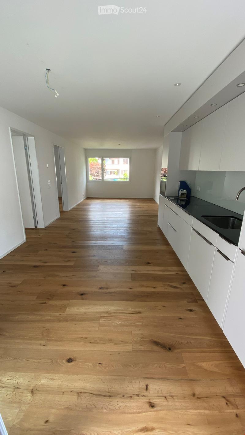 Empty room with light wooden flooring, white walls, and a small kitchen area with white cabinets and a countertop visible on the right side.