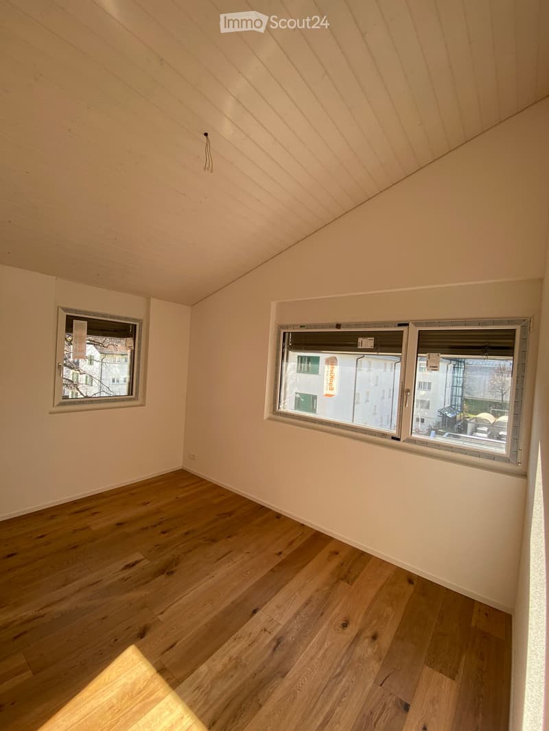 Empty room with wooden flooring, white walls, and large windows offering a view of the street.