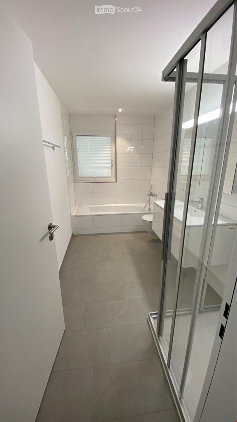Modern bathroom with a bathtub, toilet, sink, and a glass-enclosed shower area. The space features white tiled walls and a grey tiled floor.