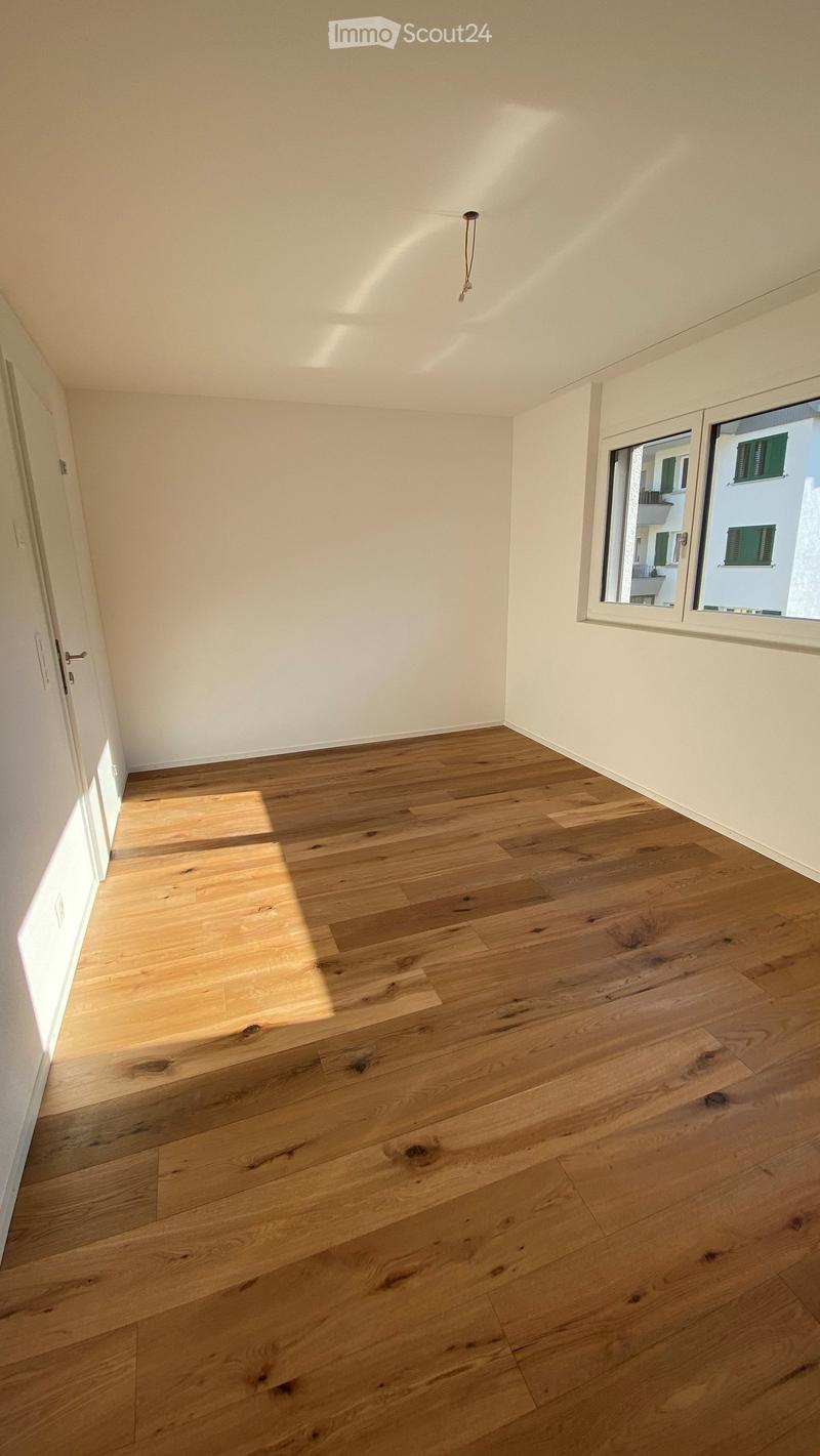 Empty room with light wooden flooring and white walls, featuring a large window with a view of neighboring buildings.