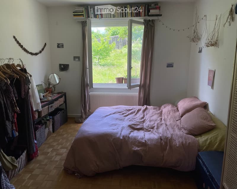 Cozy bedroom with a bed, wooden floor, large window with curtains, and a small table with a mirror and potted plant.