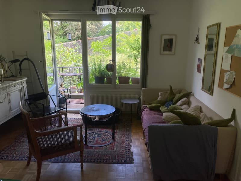 Bright living room with wooden flooring, large windows, and a balcony. Features a sofa with green cushions, a round coffee table, a wooden chair, a mirror, and a bookshelf with books.