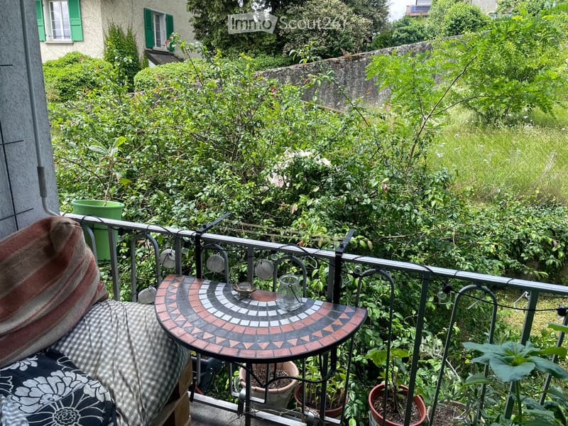 Balcony with a small round table, a chair, a cup, and several potted plants, surrounded by lush greenery and a stone wall.