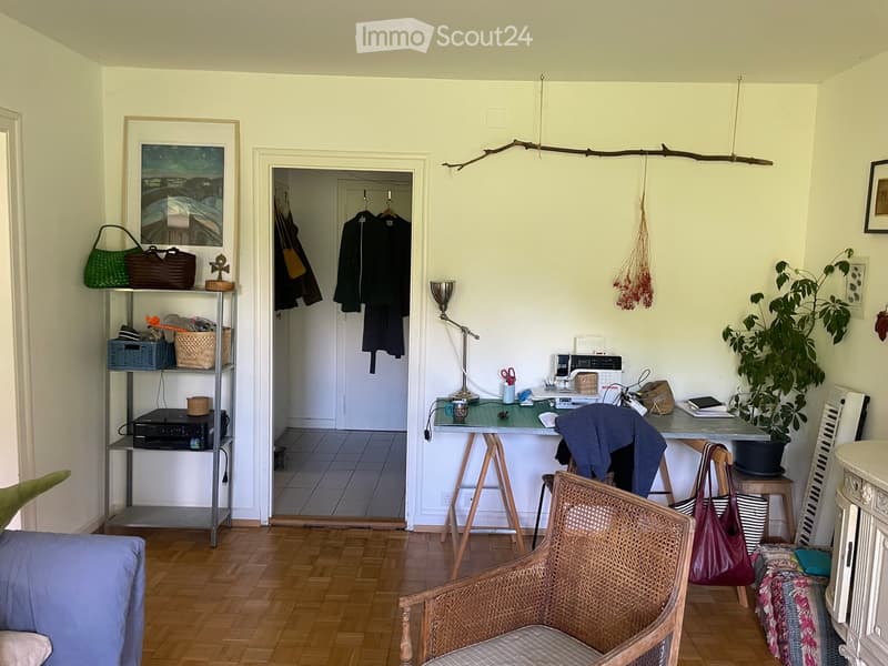 A cozy, well-furnished room with wooden flooring, a wicker chair, a small table with a lamp, a shelf with various items, and a doorway leading to another room.