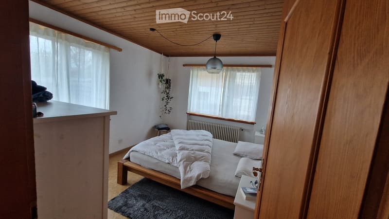 Bedroom with wooden bed, white bedding, wooden floor, white walls, wooden ceiling, white curtains, radiator, hanging plants, wooden nightstand, books, and a rug.