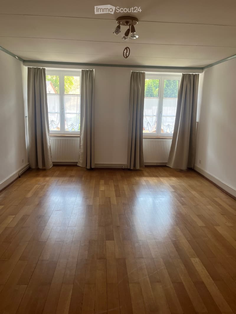 Empty room with wooden flooring, large windows with curtains, and a ceiling light fixture.