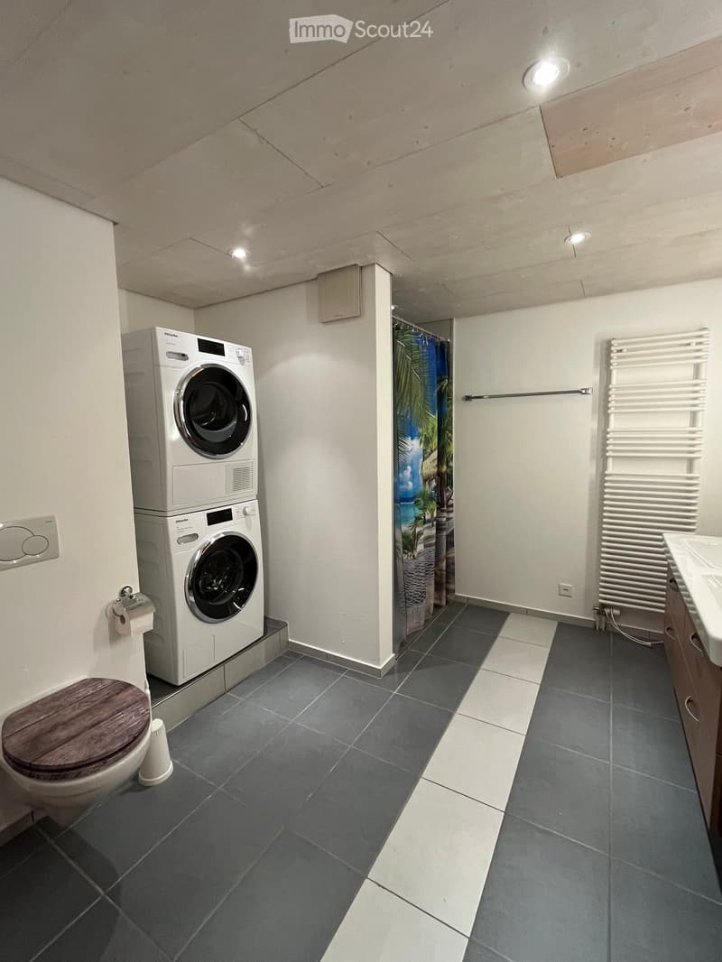 Modern bathroom with dark grey tiles, a toilet with a wooden seat, a stacked washing machine, a radiator, a sink, and a shower area with a tropical-themed curtain.
