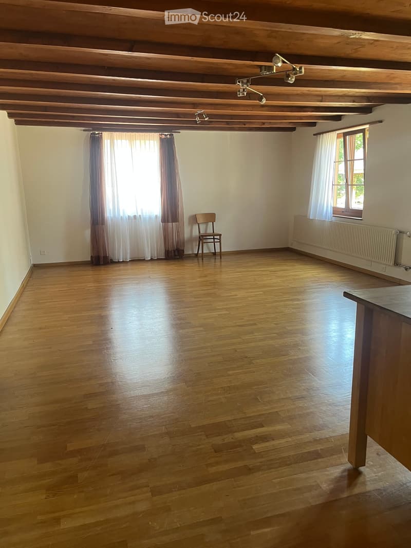 Empty room with wooden floor, exposed wooden ceiling beams, and large windows with curtains. A single wooden chair is placed near the window.