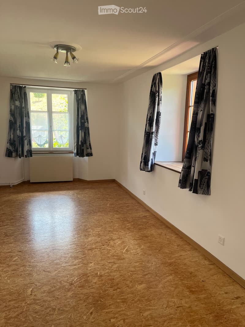 Empty room with wooden flooring, white walls, large windows with curtains, and a radiator under one window.