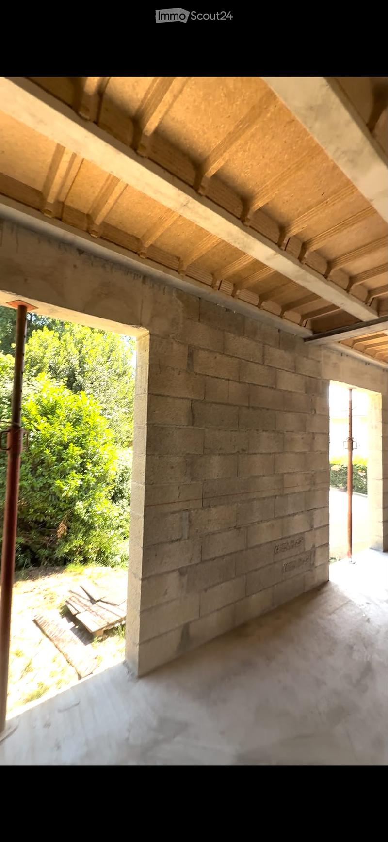 An open, unfinished corridor with exposed wooden beams and stone walls, leading to a garden area with greenery visible through an opening.
