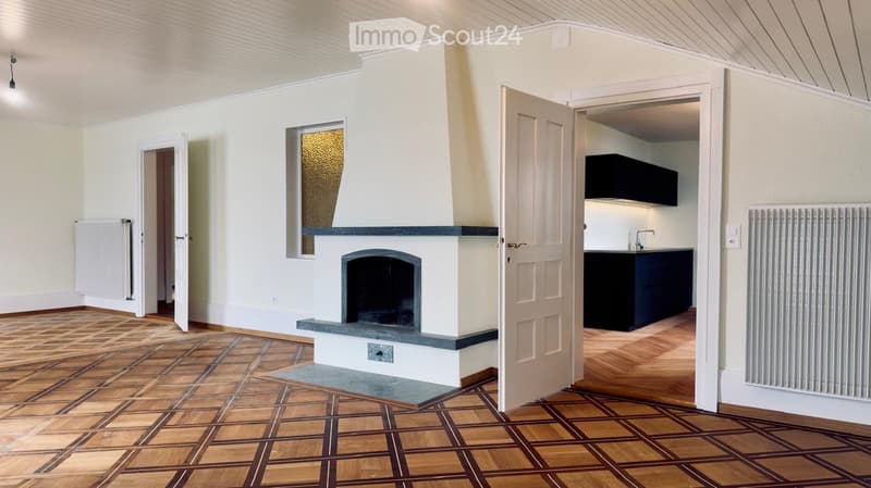Empty room with wooden flooring, white walls, fireplace, radiator, and an open doorway leading to a kitchen area.