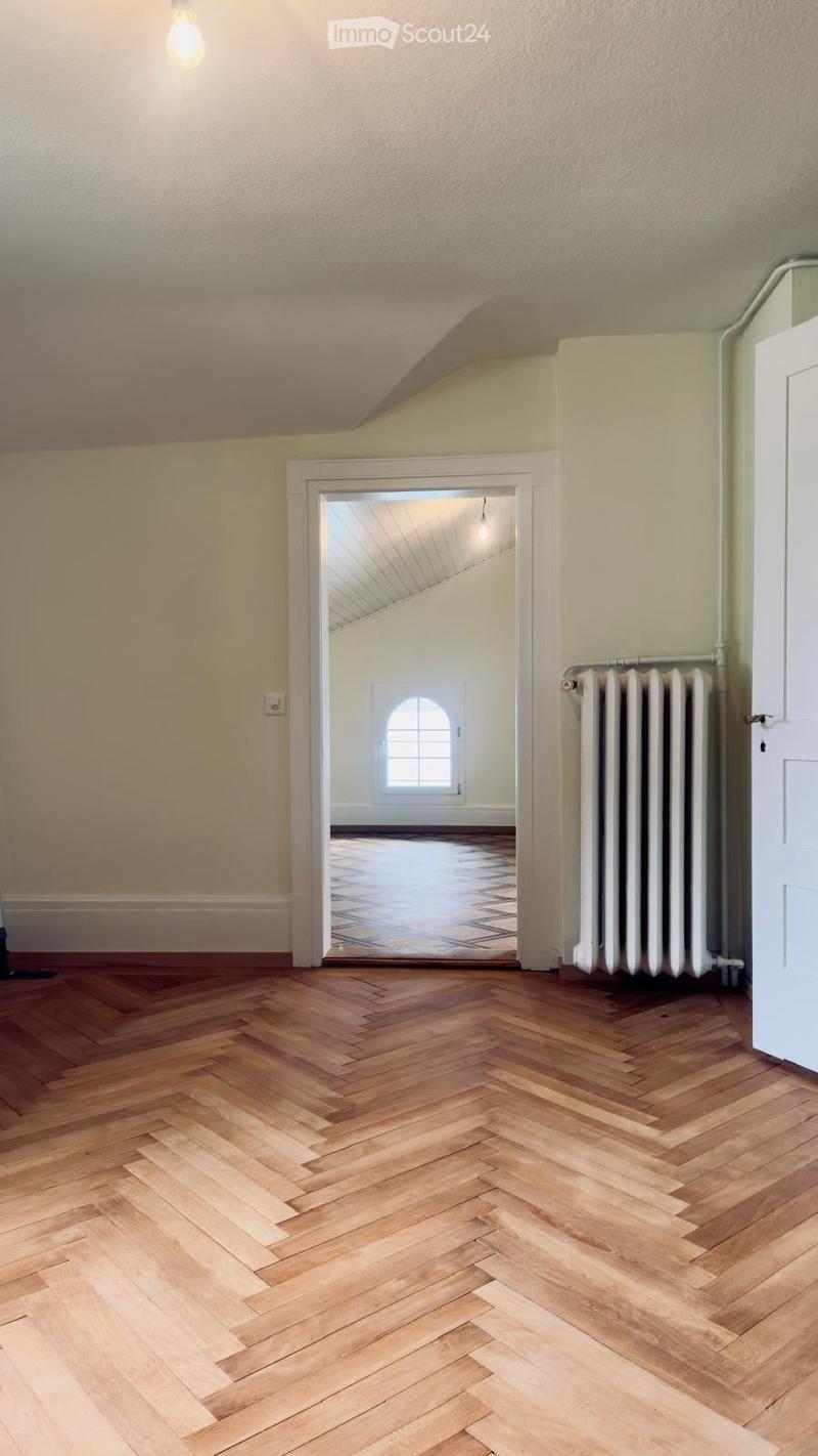 Empty room with wooden herringbone flooring, light-colored walls, and a radiator. An open doorway leads to another room with a window and patterned floor.