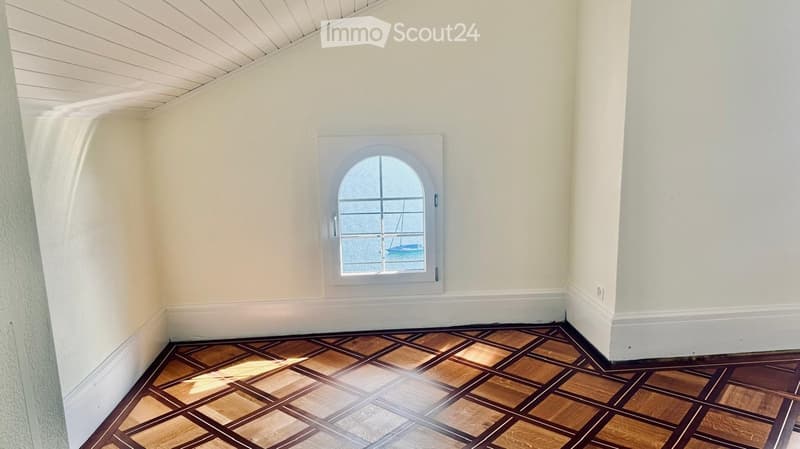 Empty room with wooden parquet flooring and white walls, featuring a small arched window with a view of a boat on the water.