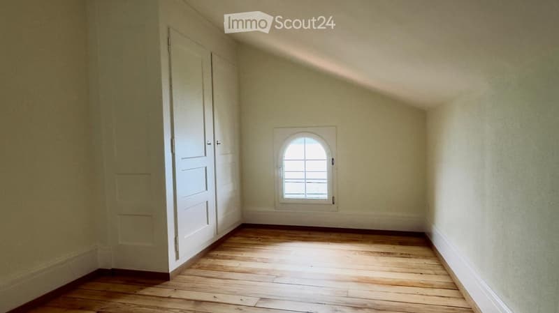 Empty room with wooden flooring, white walls, and a single arched window with multiple panes. There is a white door on the left side.