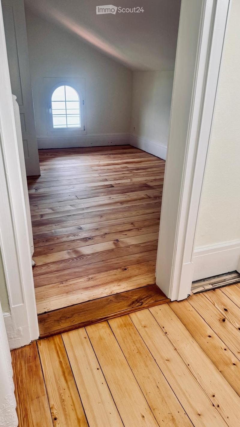 Empty room with wooden flooring and white walls, featuring a small arched window and a doorway leading to another room.