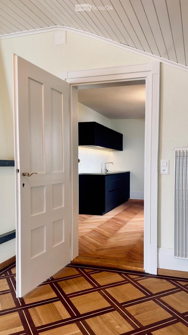 Open doorway leads to a modern kitchen with dark cabinets, a sink, and a faucet. The hallway has patterned wooden flooring and white walls.