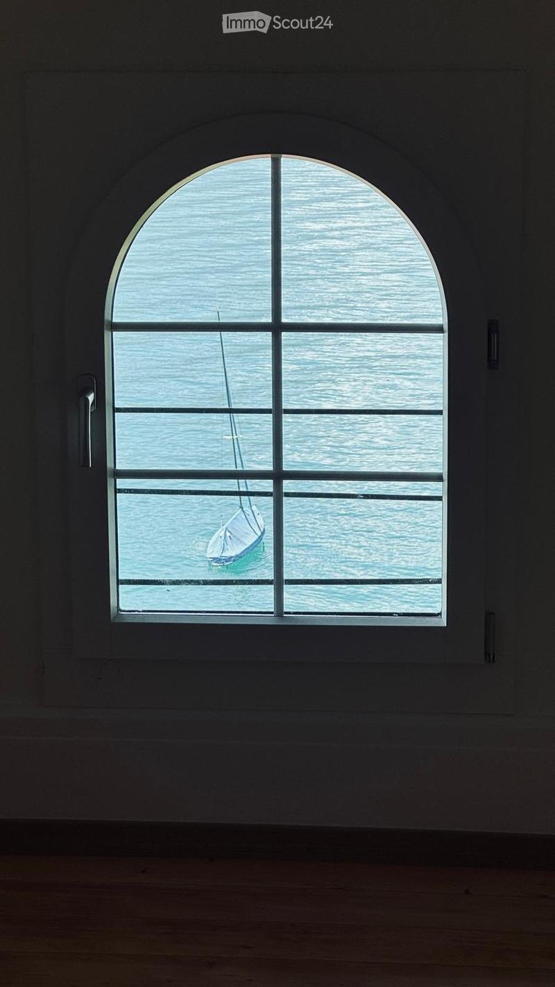 Empty room with wooden floor and large arched window showing a sailboat on the water outside.