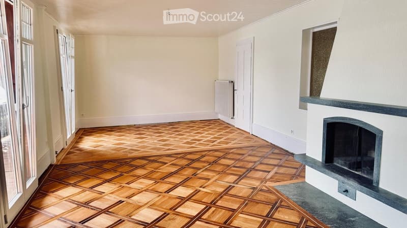 Empty room with wooden parquet flooring, white walls, fireplace, radiator, and large windows with shutters.