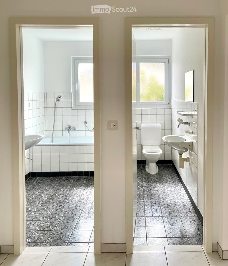 Two adjacent bathrooms with white tiled walls and grey tiled floors. The left bathroom has a bathtub with a showerhead and a sink, while the right bathroom features a toilet and a sink with a mirror above it.