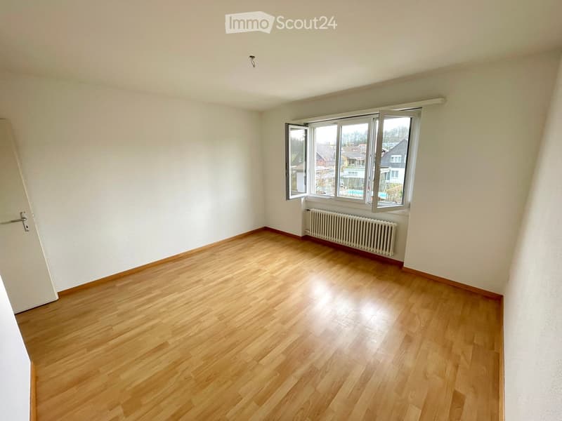 Empty room with wooden flooring, white walls, and a large window with a view of the neighborhood.