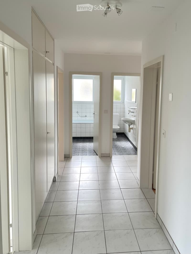 A clean, tiled hallway with white walls, leading to two bathrooms. The hallway has a central ceiling light fixture and doors on both sides.