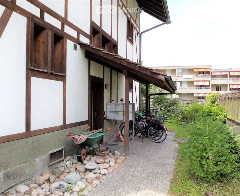 Outdoor area next to a building with a covered entrance, featuring a wheelbarrow, bicycles, and a small garden with greenery.