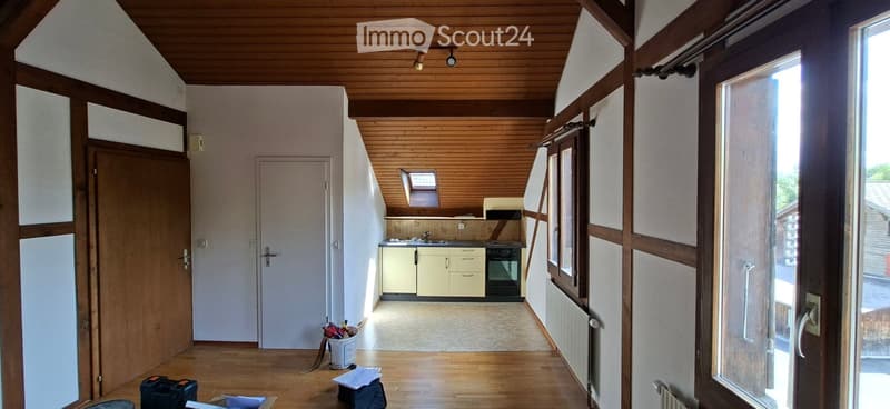 The image shows a small, well-lit kitchen area with light-colored cabinets, a stove, and a sink. The space features wooden flooring and a slanted wooden ceiling. There is a door leading to another room and a window above the sink, allowing natural light t