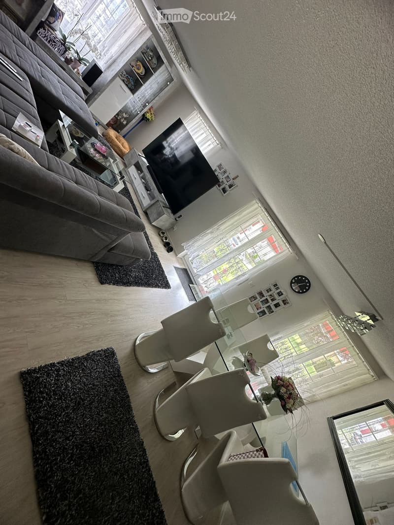 Modern living room with a grey sofa, glass coffee table, white chairs around a dining table, large windows, and a mirror on the wall.
