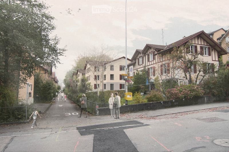 Residential street lined with multi-story houses featuring pitched roofs and varied facades, surrounded by trees and greenery. People are walking along the sidewalk.