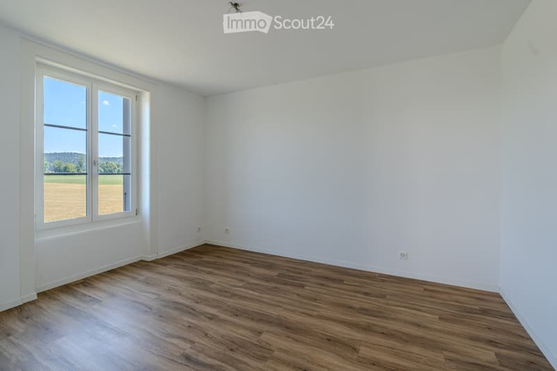 Empty room with white walls, wooden flooring, and large windows offering a view of a field and trees.