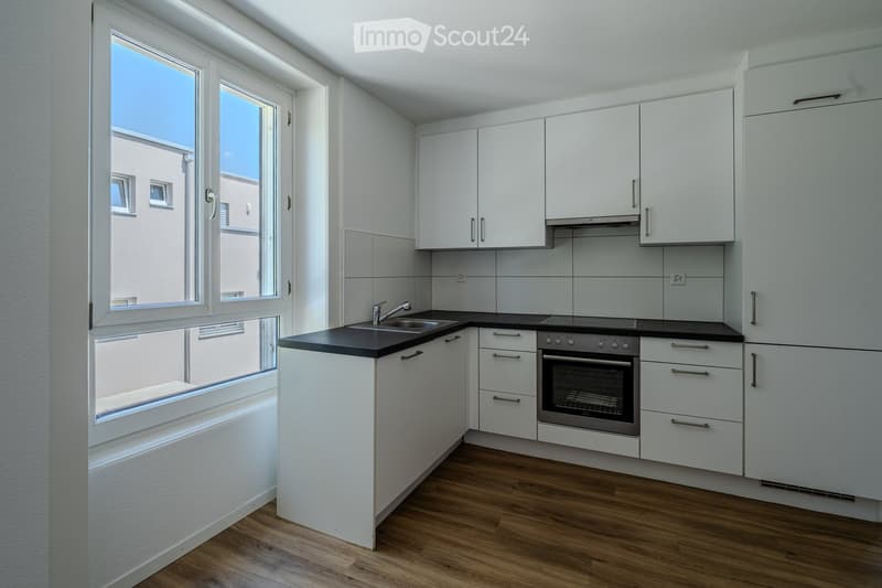 Modern kitchen with white cabinets, black countertops, a sink, faucet, and an oven. The room features wooden flooring and large windows offering a view of the outside.