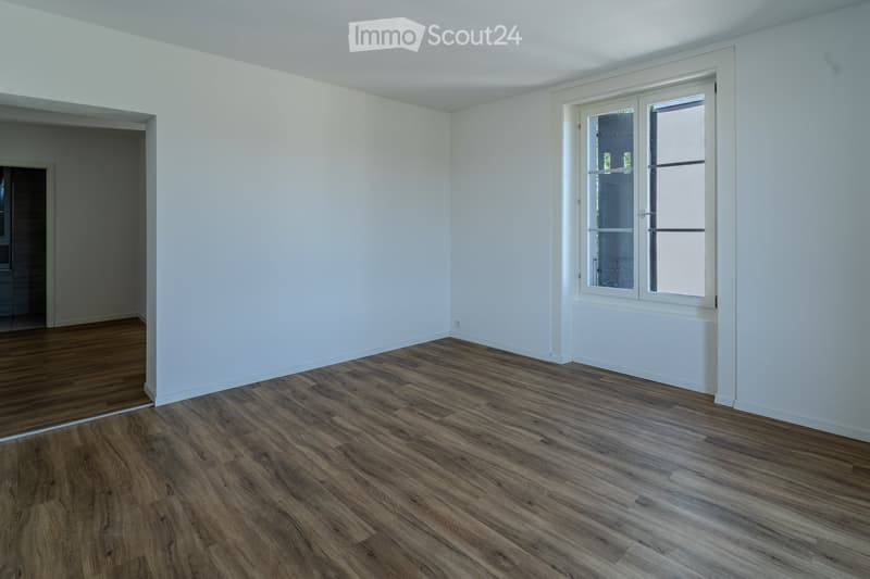 Empty room with light-colored walls, wooden flooring, and a large window with white frames. The room is unfurnished and has a neutral, minimalist design.
