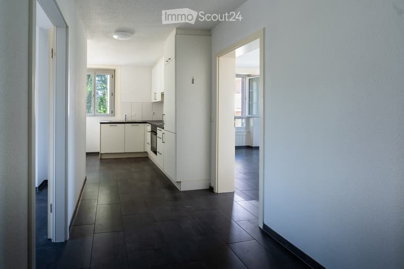 Modern kitchen with white cabinets, dark tiled floor, and a sink area. The space is well-lit with natural light from a window and has an open doorway leading to another room.