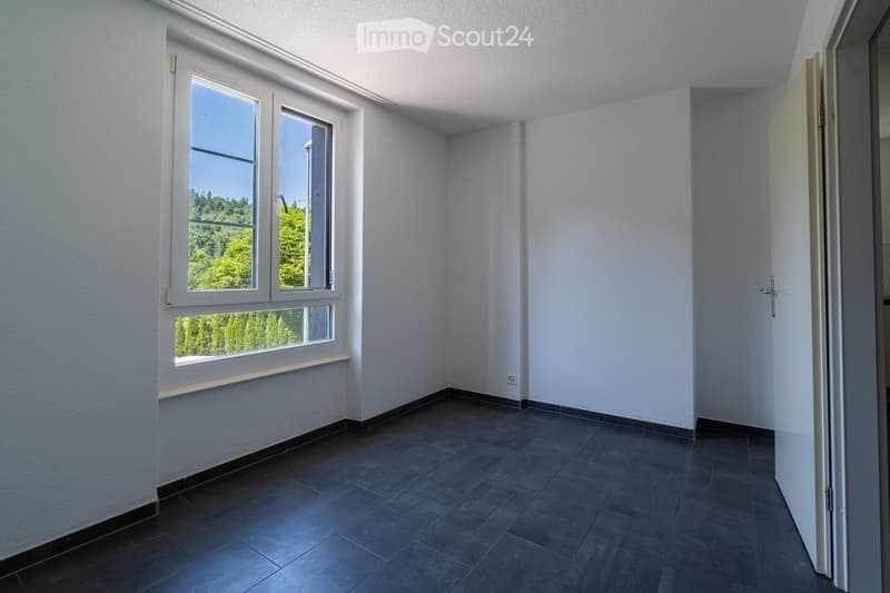Empty room with white walls, dark tiled floor, and large windows offering a view of greenery outside.