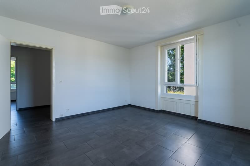 Empty room with white walls, dark tiled floor, large window with white frame, and an open doorway leading to another space.