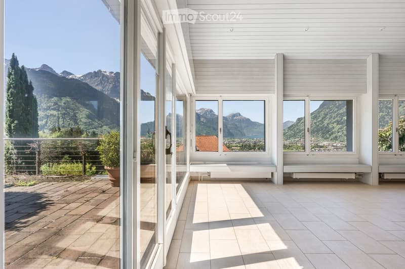 Bright, modern balcony with white wooden paneling, large windows offering panoramic mountain views, tiled floor, and built-in seating benches.