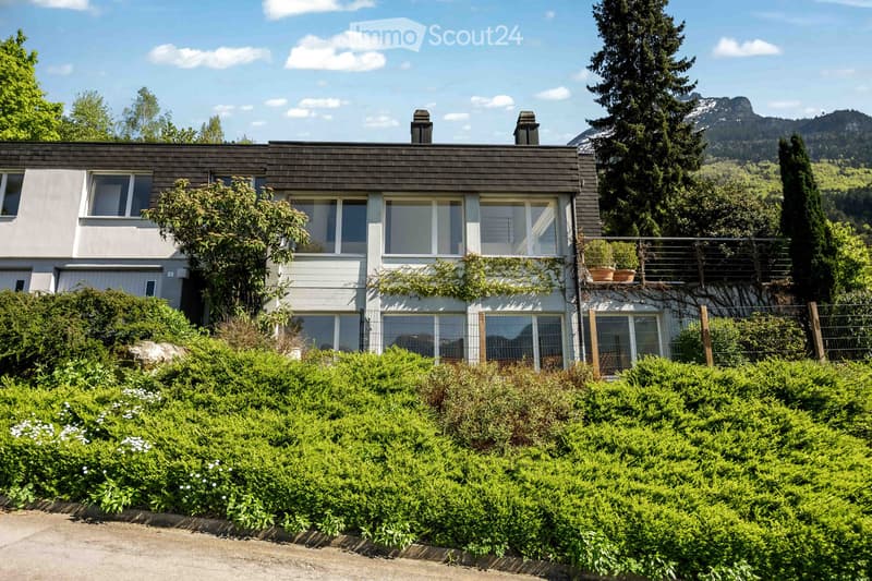 Modern two-story house with a flat roof, large windows, and a balcony on the upper level, surrounded by lush greenery and a scenic mountain backdrop.
