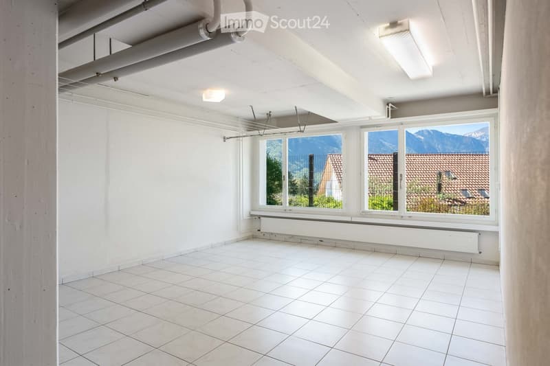 Empty room with white tiled floor, white walls, large windows showing a view of mountains and a neighboring building, and exposed ceiling pipes.