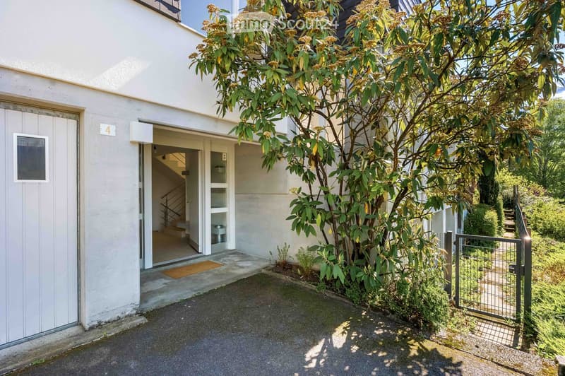 A modern white building with a small entrance area, a staircase leading up, and a tree providing shade. The entrance features a doormat and a small gate leading to a garden area.