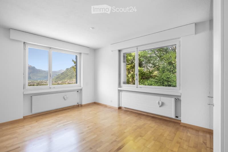Empty room with wooden flooring, white walls, large windows offering scenic views, and built-in radiators.