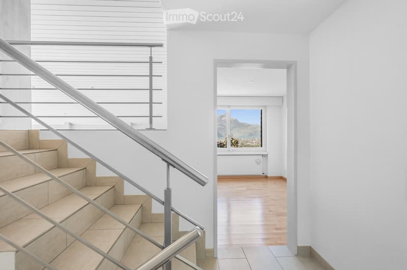 Modern hallway with a staircase featuring a metal railing and wooden steps, leading to a room with wooden flooring and large windows offering a scenic view.