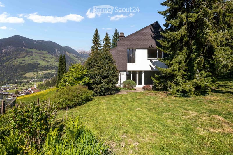 Modern two-story house with dark brown roof and white walls, surrounded by lush greenery and trees, with a scenic mountain view in the background.