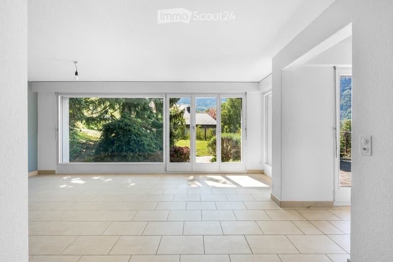 Empty room with tiled floor, white walls, large glass doors leading to a garden, and a single light fixture on the ceiling.